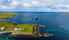 A large blue ship sails near the rocky coastline of a rural area with green fields and scattered buildings under a partly cloudy sky.