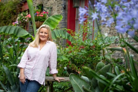 A person with long blonde hair leans on a wooden railing in a garden with lush greenery and purple flowers. A brick building with a red door and hanging plants is in the background.