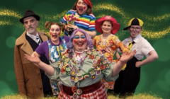 A group of six individuals in colorful costumes pose cheerfully in front of a green background with glittery accents. The text reads "Open Door Drama's Babes in the Wood.