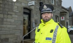 A police officer in a bright yellow uniform stands outside a stone police station building.