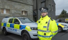 Police officer in a high-visibility jacket stands by two police vehicles outside a stone building.