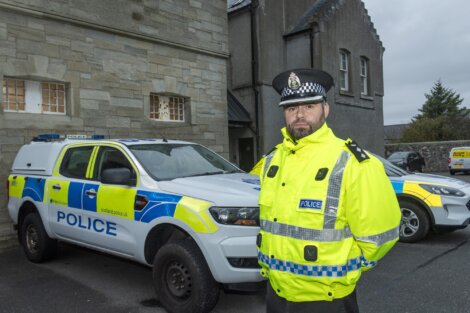 Police officer in a high-visibility jacket stands by two police vehicles outside a stone building.