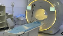 MRI machine in a hospital room, with adjustable bed positioned for patient scanning. Monitors display settings. Room contains medical equipment and supplies.