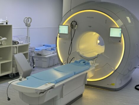 MRI machine in a hospital room, with adjustable bed positioned for patient scanning. Monitors display settings. Room contains medical equipment and supplies.