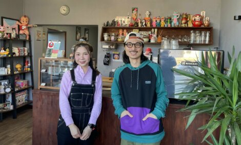 Two people standing in a cozy cafe adorned with toys and collectibles, in front of a counter with a coffee machine. A potted plant is on the right.