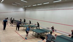 People playing table tennis in a large indoor room with multiple tables.