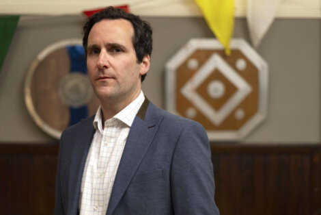 A man in a blue blazer and white shirt stands indoors, with decorative shields on the wall behind him.