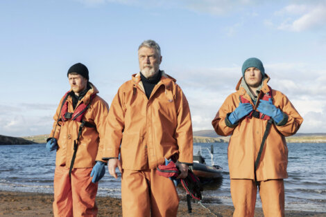 Three people in orange raincoats and life vests stand on a beach, with a boat and sea in the background.