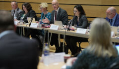 A group of people sit at a long table with laptops and microphones during a meeting in a conference room.