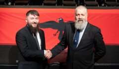 Two men in suits with beards shake hands in front of a red flag featuring a black raven.