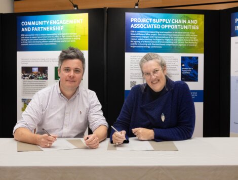 Two people sitting and signing documents at a table with project information boards in the background.