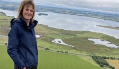 A person stands on a hill overlooking a large lake and green fields under a cloudy sky.