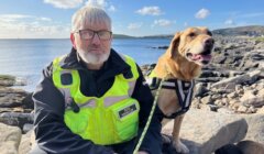 A man wearing a high-visibility vest sits on rocks by the sea with a dog on a leash.