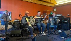 Four musicians perform with accordions, a piano, and drums on a stage with red and gold decor.