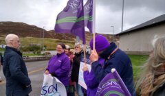 A group of people holding purple flags and signs stands outside on a cloudy day, engaging in conversation.