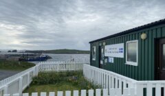 Small ferry terminal building by the sea, with a white fence in the foreground and boats moored in the background under a cloudy sky.