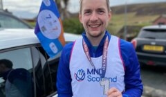 A man smiling, wearing a Mind Scotland bib, holding a medal with the number "1" on it. There's a Mind Scotland flag attached to a nearby car in the background.