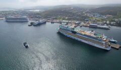 Aerial view of a harbor with three large cruise ships docked and a smaller boat navigating the water. Buildings and hills are visible in the background.