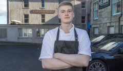 Young chef in a white jacket and dark apron stands with arms crossed outside a stone building with a sign reading "The Garret".