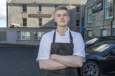 Young chef in a white jacket and dark apron stands with arms crossed outside a stone building with a sign reading "The Garret".