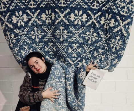 A person stands in front of a large blue and white patterned textile, holding a small sign that reads "Dundee Design Festival.