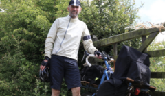 Man in cycling gear stands beside a loaded bicycle near greenery.