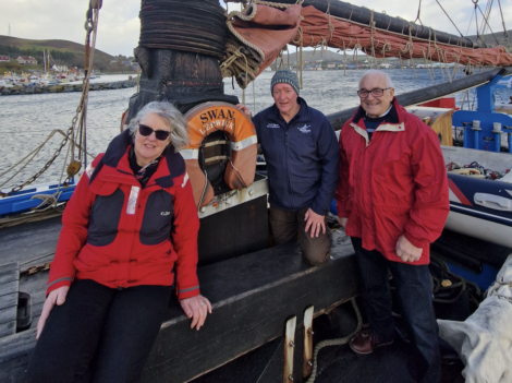 Three people on a boat deck with a life preserver labeled "Swan Lerwick" in the background. The water and distant shore are visible.