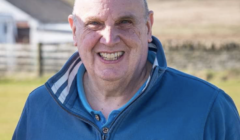 A man in a blue jacket smiles outdoors with blurred buildings and fields in the background.