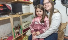 A woman and a young girl sit in a room with shelves containing pet supplies. A cat is visible in a cage on the shelf.