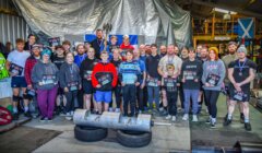 A group of people poses with awards in a gym, featuring weightlifting equipment in the foreground and a Scottish flag on the wall.