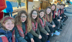 Nine children wearing life vests sit in a row on a boat deck, smiling and facing the camera.