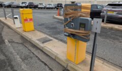 A parking ticket machine is wrapped with brown tape, next to a yellow waste bin. Several parked cars are in the background.