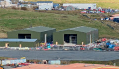 Two green industrial warehouses are under construction, surrounded by building materials and machinery on a hillside with additional buildings and parked cars in the background.