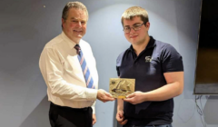 Two men stand indoors; one in a white shirt and tie, the other in a navy polo. They are jointly holding a rectangular engraved plaque.