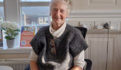 An elderly person with white hair sits in an office chair, wearing a gray knitted vest over a cream turtleneck. A potted plant and sign are on the windowsill in the background.