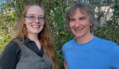 Two people are standing outdoors in front of greenery, smiling at the camera. The person on the left has long red hair and glasses, and the person on the right has short gray hair and is wearing a blue shirt.