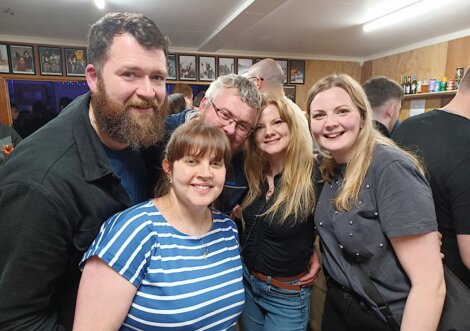 Five people smiling and posing together in a crowded room with framed pictures on the walls.