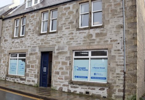 A stone building houses Lerwick Dental Practice, featuring windows with blue and white signage. An entrance sign is visible beside a blue door.