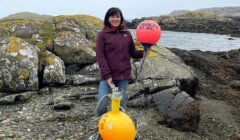 A woman stands on a rocky shore holding two buoys, one orange and marked "1023" and the other red and marked "SO 763." A small body of water is visible in the background.