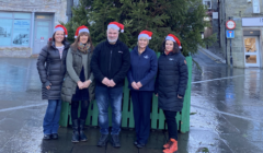 Five people in winter clothing and Santa hats stand in front of a decorated tree in a wet cobblestone square.