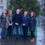 Five people in winter clothing and Santa hats stand in front of a decorated tree in a wet cobblestone square.