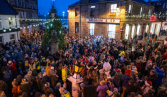 A large crowd gathers in a festive town square at dusk, surrounded by buildings and holiday lights.