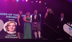 Group of people on stage at Scotland's Health Awards, two women holding a trophy. A photo of Lucy Flaws, NHS Shetland, "Leader of the Year" winner, is displayed on a screen.