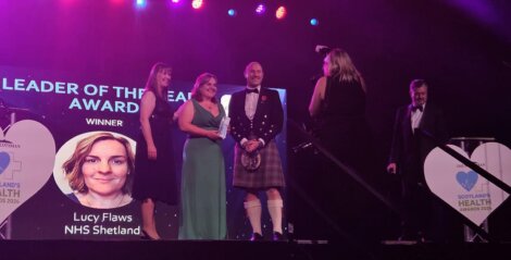 Group of people on stage at Scotland's Health Awards, two women holding a trophy. A photo of Lucy Flaws, NHS Shetland, "Leader of the Year" winner, is displayed on a screen.