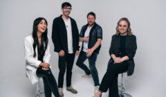 Four people pose together in a studio. Two are seated on stools, while the other two stand. The background is plain white.