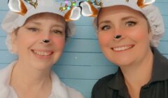 Two people wearing white caps with a deer filter, including antlers and ears, on their heads. They are smiling in front of a blue background.