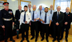 A group of ten men, some in uniforms and others in suits, are standing indoors. Four men in blue shirts are holding medals.