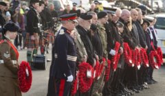 People in uniform and others stand in a line holding wreaths during a remembrance ceremony. A bagpiper is visible in the background.