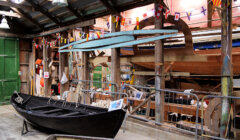 Wooden workshop interior with boats in progress, colorful flags above, and tools scattered around. A black boat is in the foreground, and a blue canoe is suspended from the rafters.