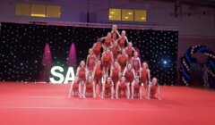 A group of cheerleaders performs a pyramid formation on a red floor with a backdrop of lights and balloons.